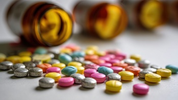 A variety of colorful pills and tablets are scattered on a white surface. Brown glass bottles are slightly blurred in the background, suggesting a recent use or examination of medication.